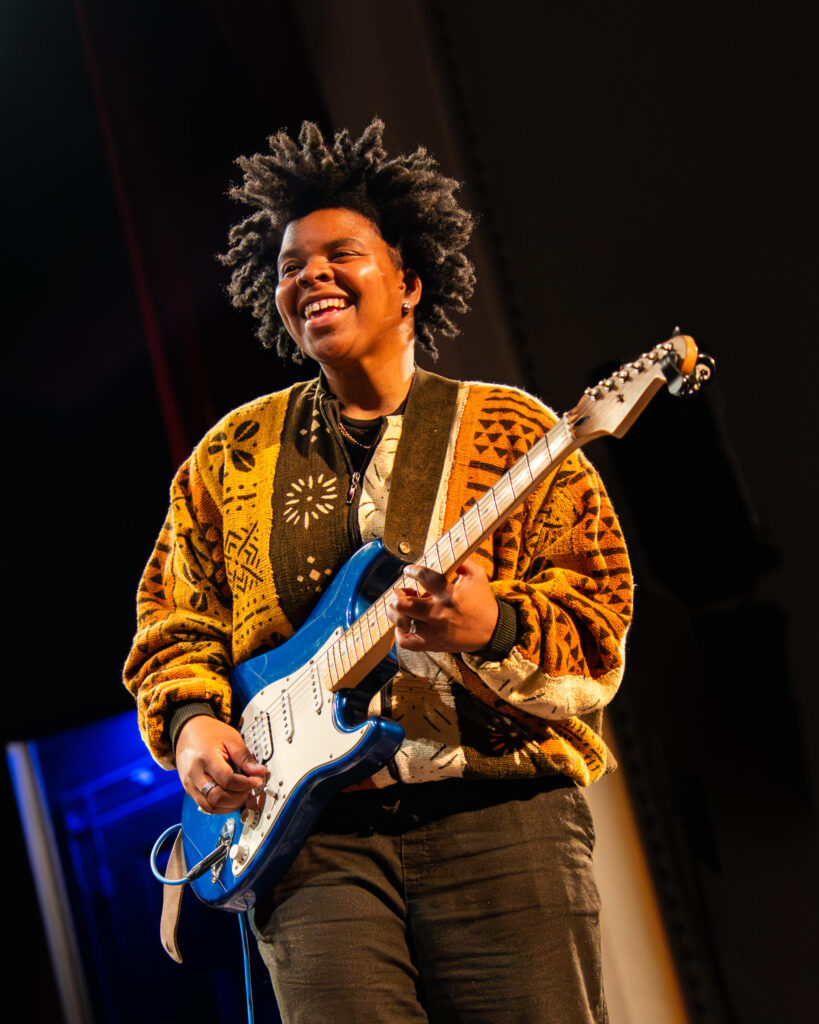 A person with dark hair and skin wearing a gold and brown patterned sweater plays a blue guitar.