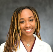 Photo of a smiling young black woman with long braids in a white medical coat.
