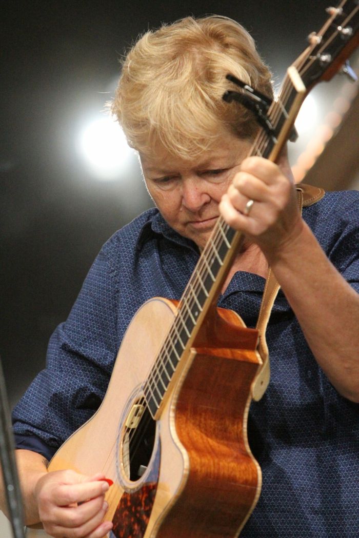 A guitarist in a navy shirt looks down while playing an acoustic guitar with a capo, lit by a spotlight on a dark stage.