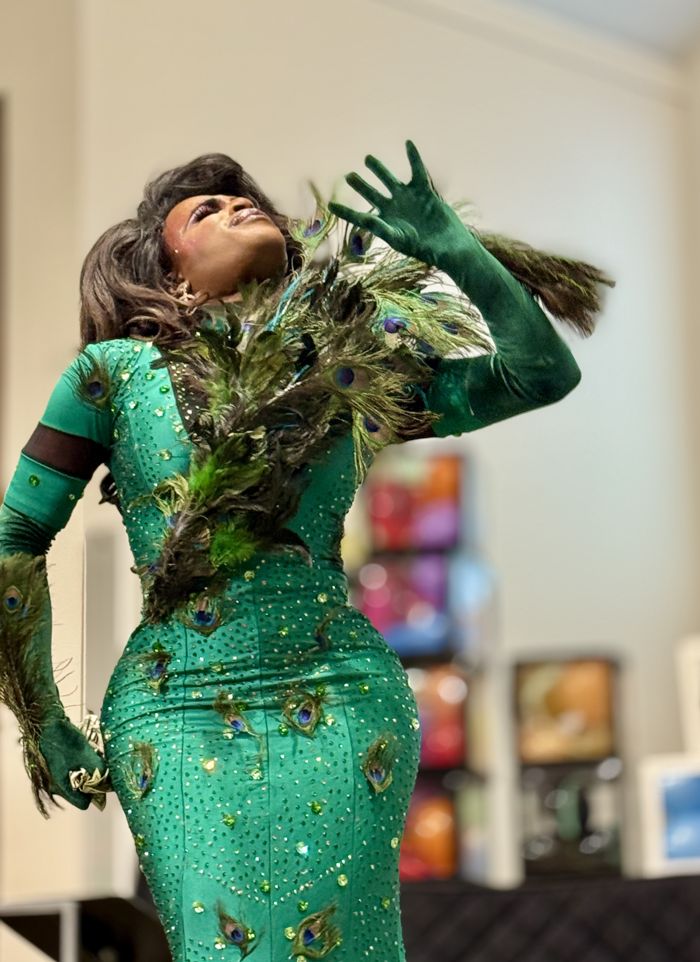 A drag performer poses in an emerald sequined gown trimmed with peacock feathers.