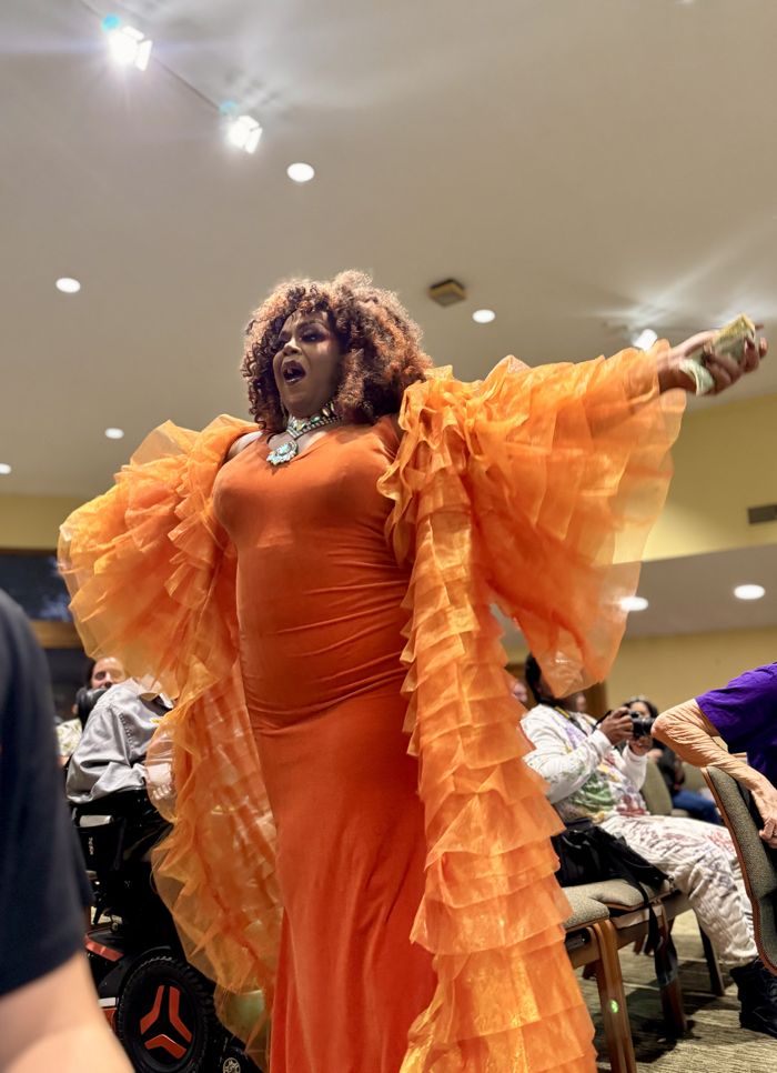 A drag performer in a huge orange ruffled gown spreads their arms and sings to the audience.