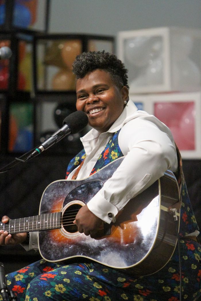 A performer in a white shirt and floral vest smiles wide while playing an acoustic guitar at the mic.