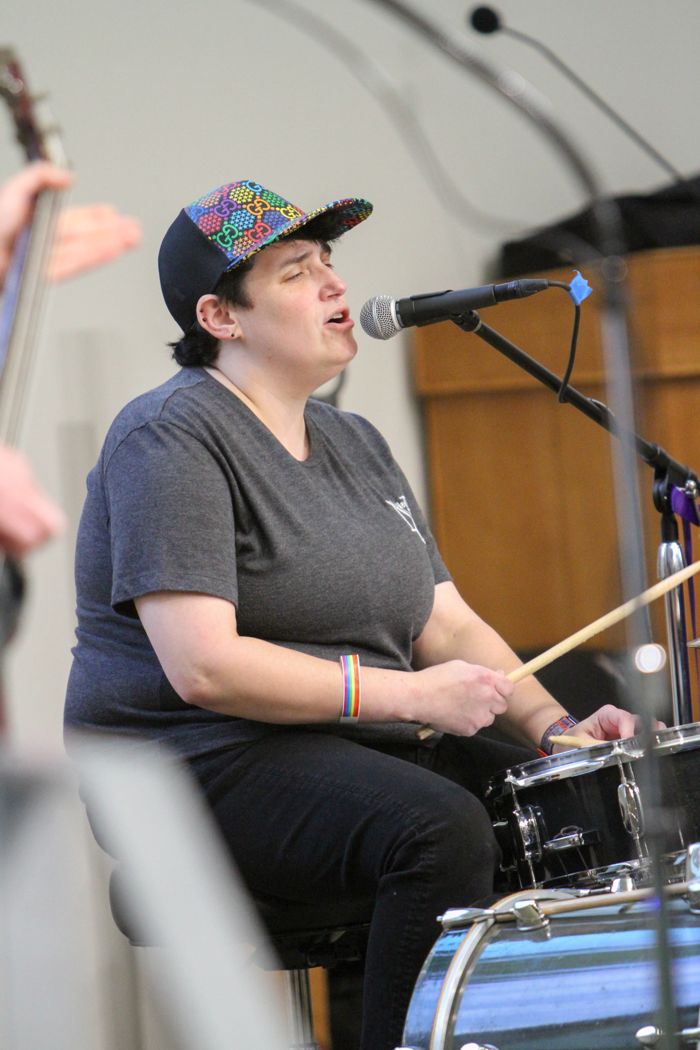 A drummer in a rainbow-print cap sings into a microphone while playing.
