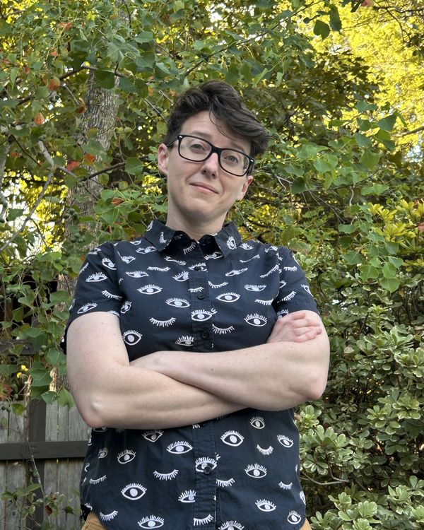 Carrie Eicherson, arms crossed with a knowing half-smile, in black glasses and a black shirt printed with eyes, outdoors among green leaves.
