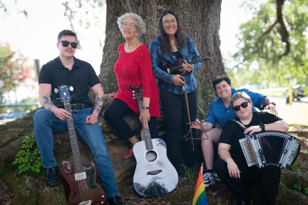 The five members of the Gais Do Do Band sit with their instruments in front of a large oak tree, a small rainbow flag at their feet.