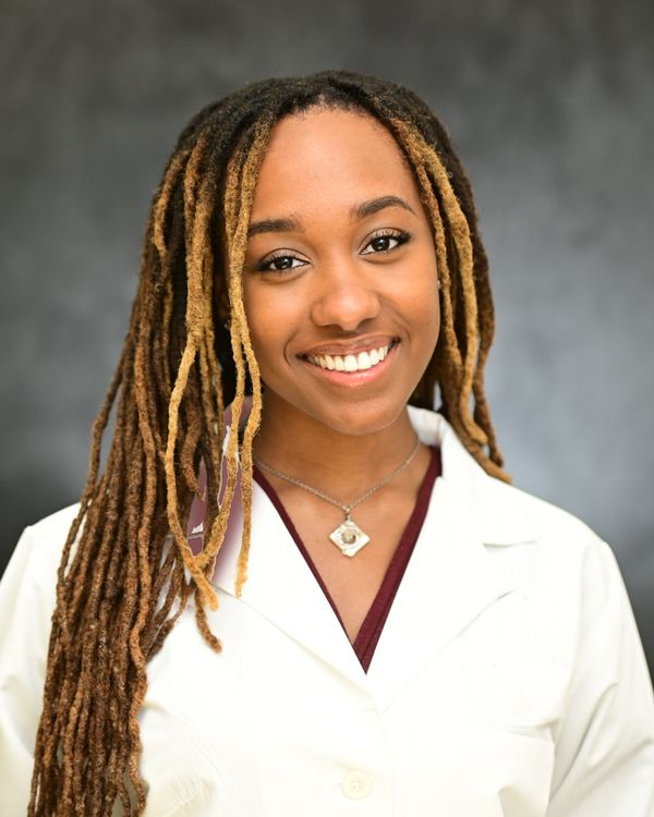 Jaidyn Bryant, smiling, with long braids, in a white coat over a maroon top, against a gray backdrop.