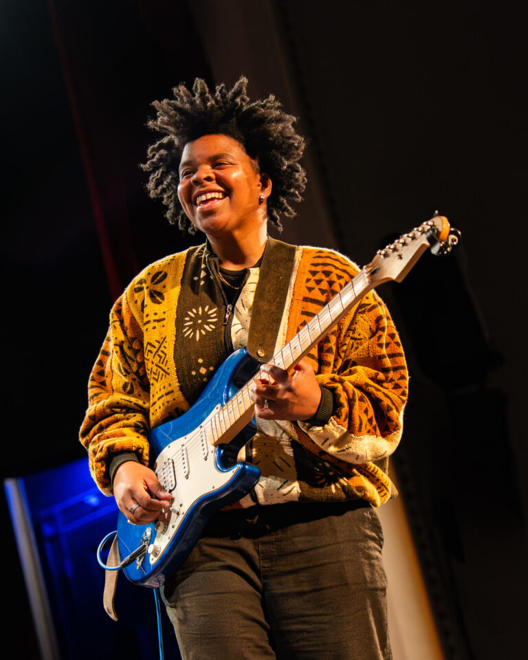 Joy Clark, smiling, plays a blue electric guitar on stage in a gold and brown patterned sweater.
