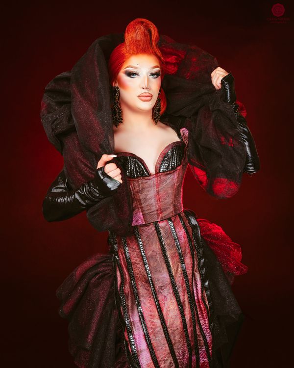 Kozmik, a drag performer with sculpted red hair, in a structured black and red gown against a deep red backdrop.