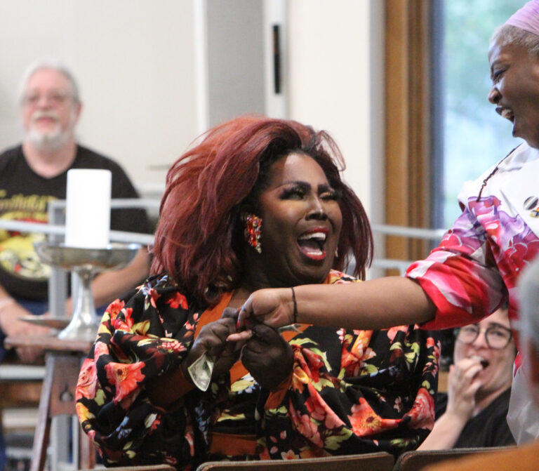 A performer with red hair and a floral top laughs and reaches toward the crowd at QUUeer Fête 2025.