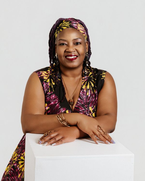 Tekoah Boatner, smiling, in a purple and yellow patterned headwrap and dress, arms folded on a white pedestal.