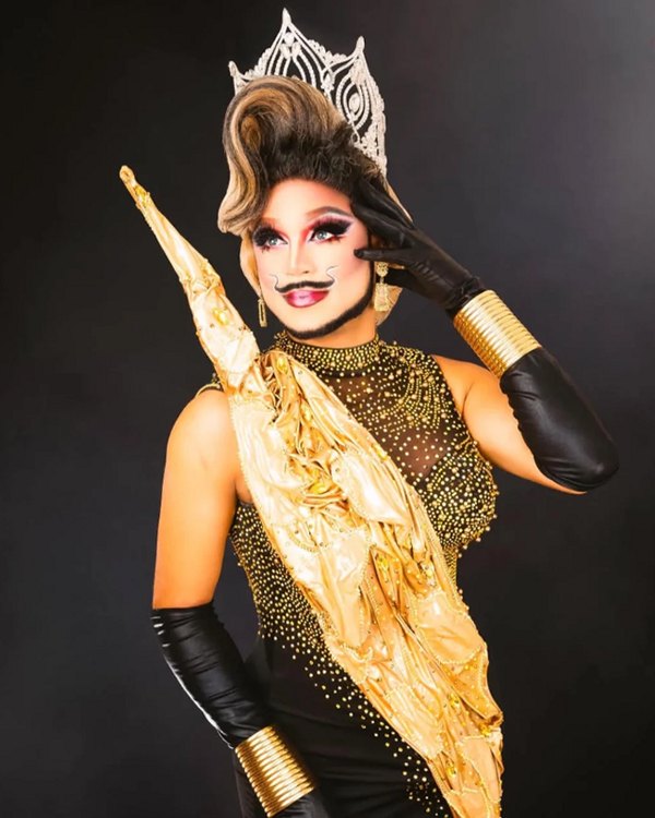 Vantasia Divine, a drag performer in a rhinestone crown and drawn-on mustache, wearing a gold-draped black gown and long black gloves against a dark backdrop.